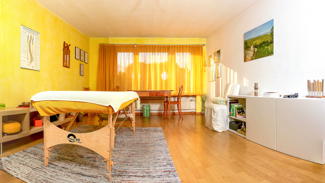 Das Bild zeigt einen hellen, sonnendurchfluteten Behandlungsraum, der wohl für alternative Heilmethoden genutzt wird. Die Wände sind in einem warmen Gelbton gestrichen. Im Raum steht eine Massageliege im Vordergrund, daneben ein kleiner Schrank und ein Teppich. Im mittleren Hintergrund befindet sich ein Schreibtisch mit Stuhl und an der Wand ein Bild mit einer Landschaft. Weitere Bilder an den Wänden und ein Regal mit Büchern und Gegenständen deuten auf einen Praxisraum hin. Die allgemeine Atmosphäre wirkt ruhig und entspannend.