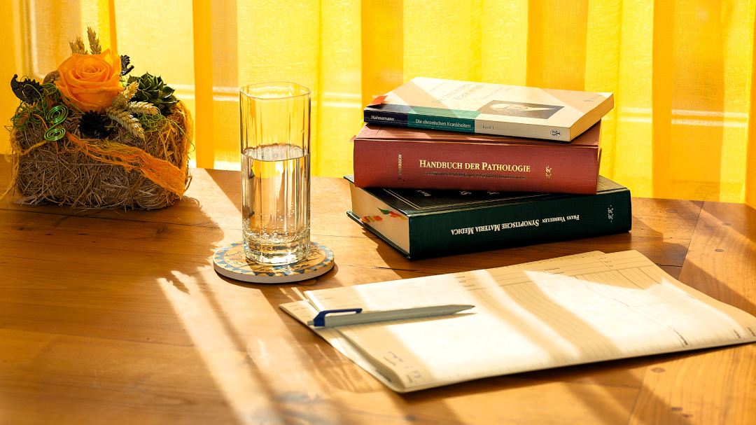 Das Bild zeigt einen hellen Holztisch, auf dem sich ein Glas Wasser, ein kleiner Blumenstrauß in einem Strohkörbchen, ein Stapel Bücher und ein Notizblock mit einem Kugelschreiber befinden. Die Bücher scheinen medizinischen Inhalts zu sein. Das Licht fällt von einem Fenster mit gelben Vorhängen auf den Tisch. Die Szene wirkt ruhig und geordnet, fast wie in einem Arzt- oder Therapiezimmer.